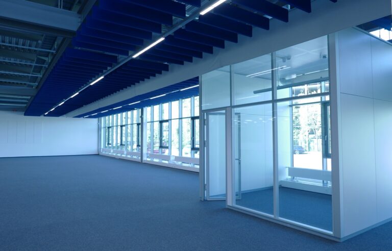 Moderner leerer Raum mit großen Fenstern, blauen Akustikelementen an der Decke und einem verglasten Bürobereich.