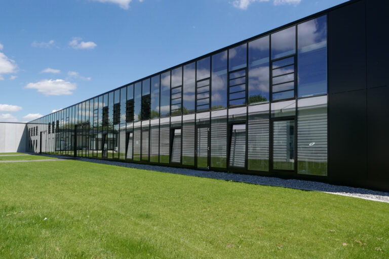 Modernes Industriegebäude mit Glasfassade, reflektierenden Fenstern und grüner Rasenfläche.