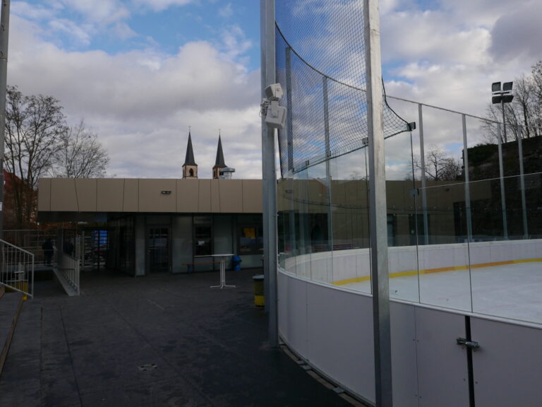 Eine Außenansicht der Eislaufbahn Würzburg mit Glasbarrieren, im Hintergrund ein modernes Gebäude und Kirchtürme.