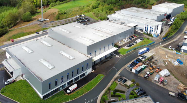 Luftbild eines Industriegebiets mit großen Lagerhallen, flachen Dächern, parkenden Fahrzeugen und umgebender Waldlandschaft.