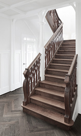 Elegante dunkle Holztreppe mit kunstvoll geschnitzten Geländern, umgeben von weißen Wänden und Decke, sowie einem dunklen Parkettboden im Fischgrätmuster.